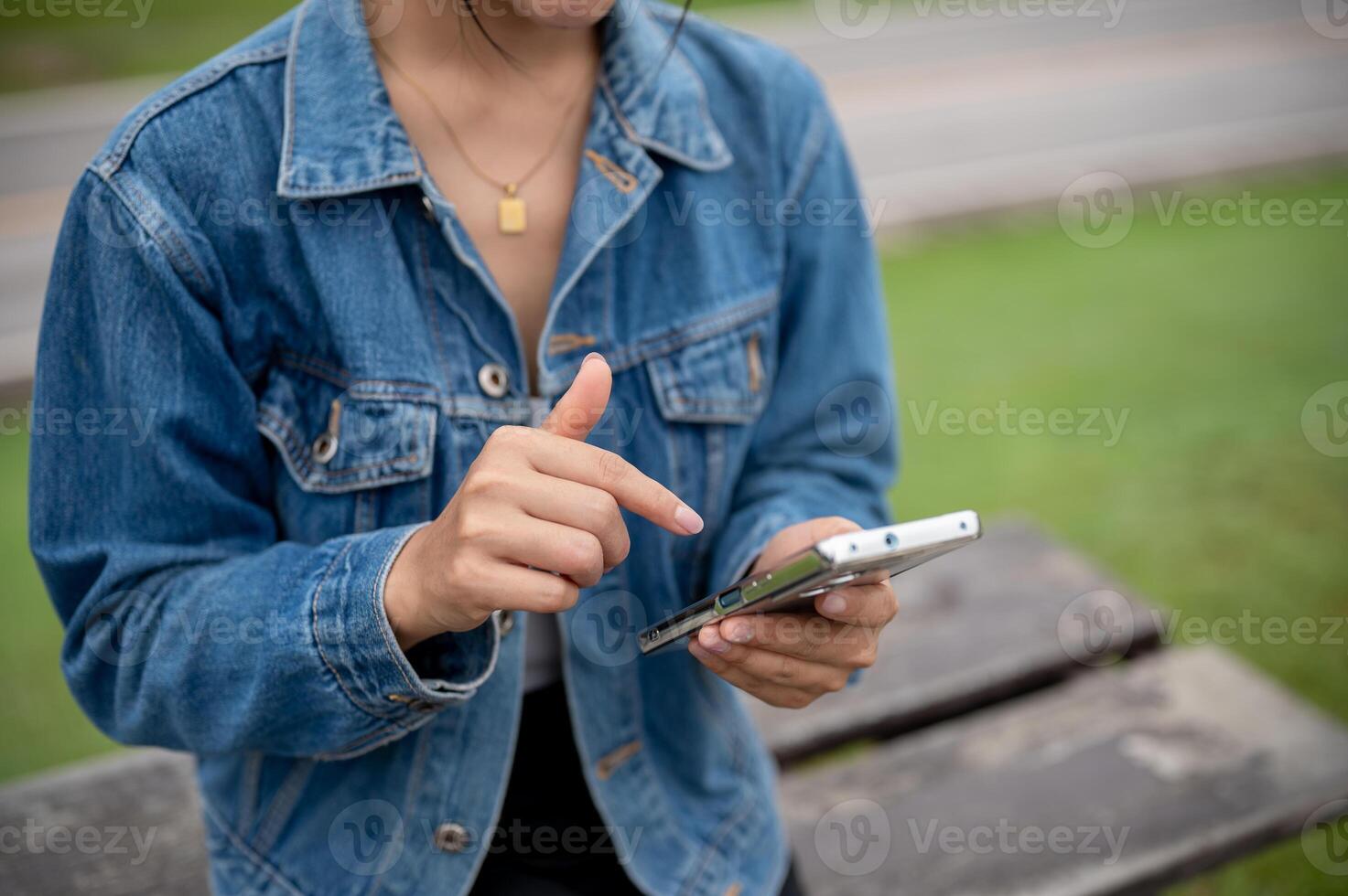 A cropped shot of a woman in a denim jacket using her smartphone while sitting on a bench in a park. photo