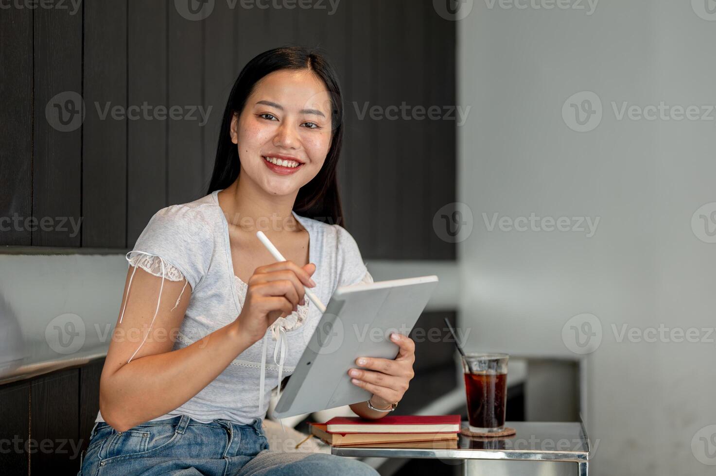 A beautiful Asian woman using a tablet, working remotely from a coffee shop, smiling at the camera. photo