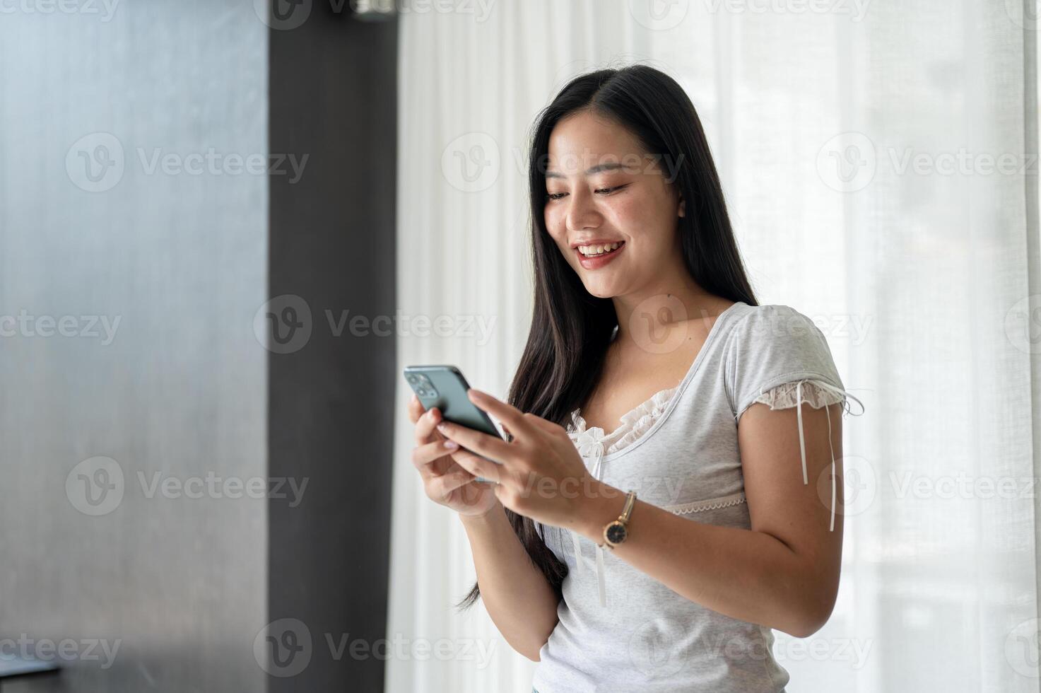 A charming Asian woman is smiling at text messages on her phone while standing by the window. photo
