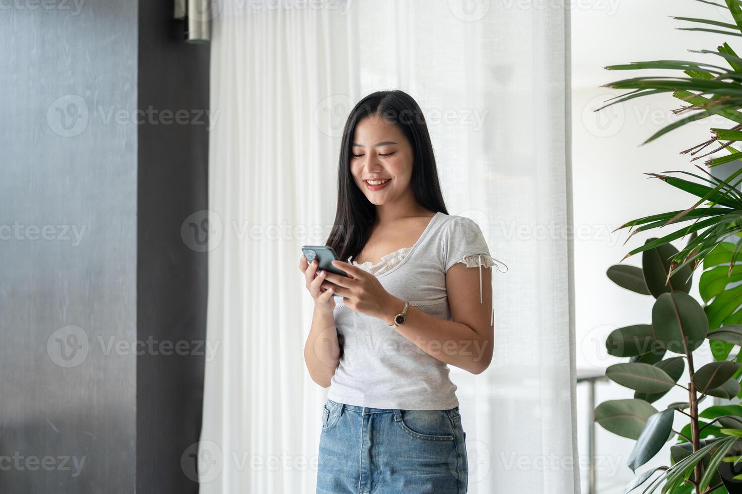 A charming Asian woman is smiling at text messages on her phone while standing by the window. photo