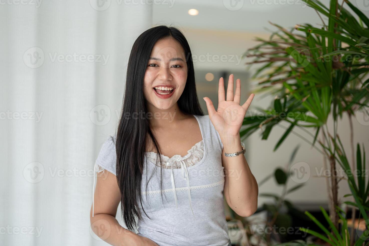 A cheerful Asian woman is standing by a window in a room, smiling and waving her hand at the camera. photo