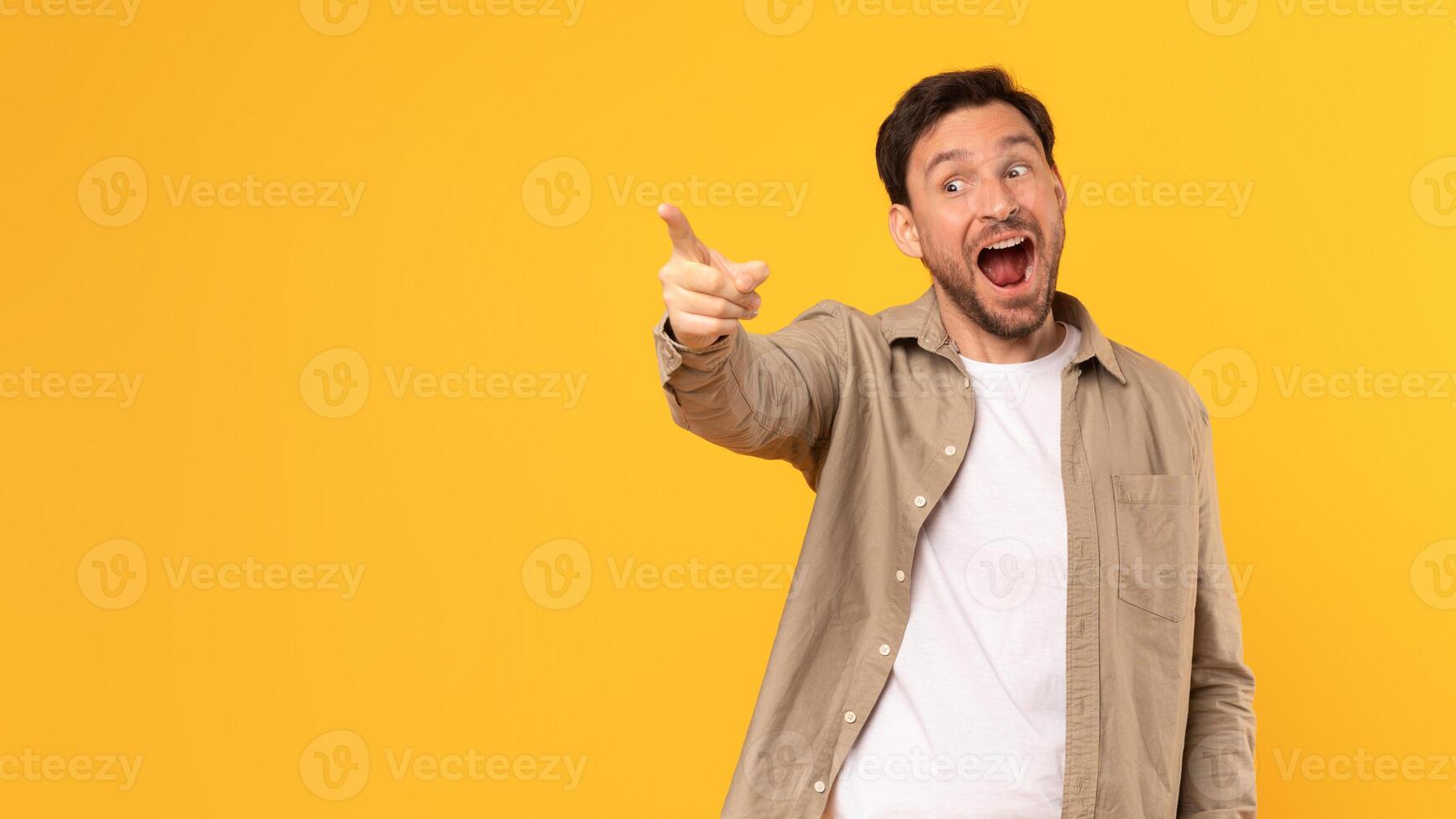 A man in a brown shirt stands in front of a yellow background, pointing his index finger at copy space and looking excitedly in that direction, with his mouth open in a smile. photo