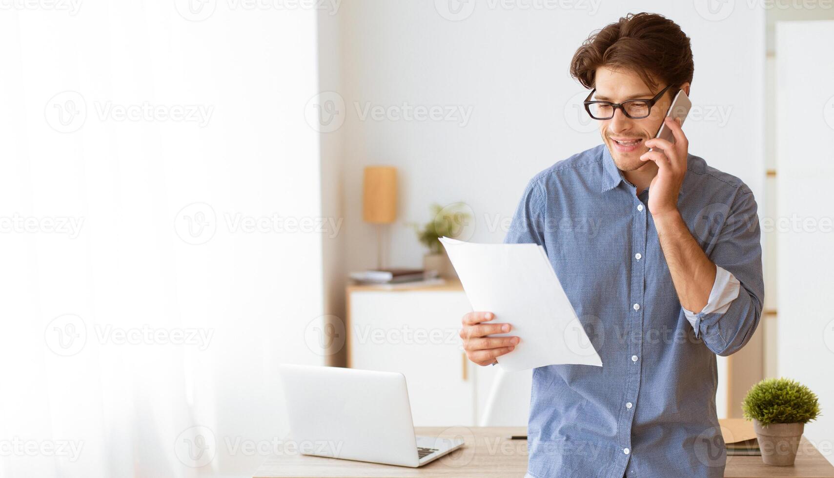 A man in glasses stands at his desk, talking on his phone while reviewing documents. photo