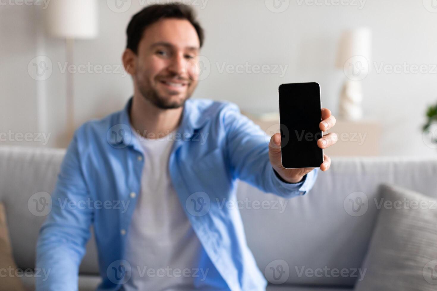 A cheerful man in a blue shirt is extending his arm to proudly display a smartphone with a blank screen, sitting on a sofa in a well-lit, cozy living room environment during the day. photo