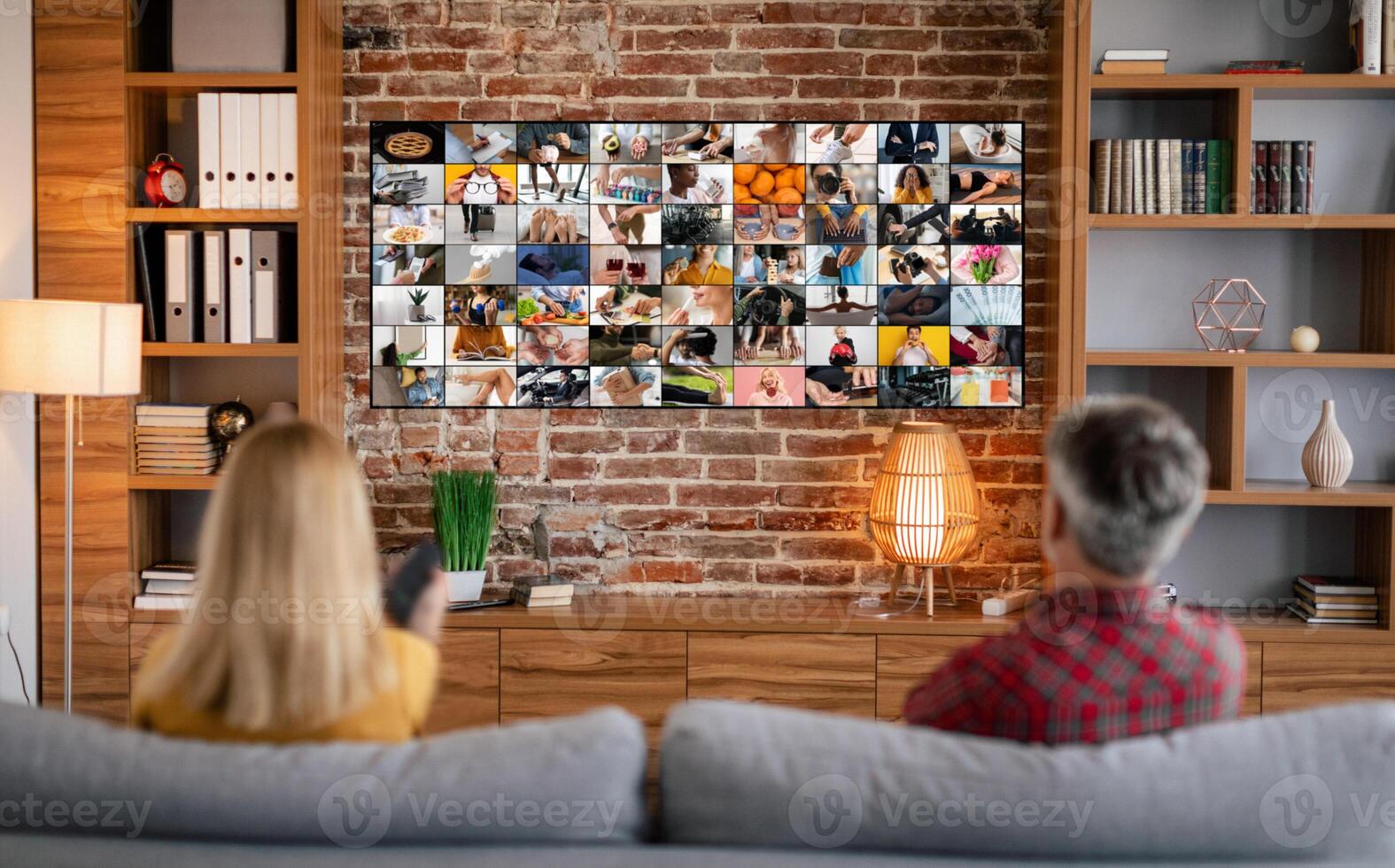 A couple relaxes on a couch in their living room, facing a large television screen displaying a grid of various images. The room features wooden shelves filled with books and decor, a brick wall photo