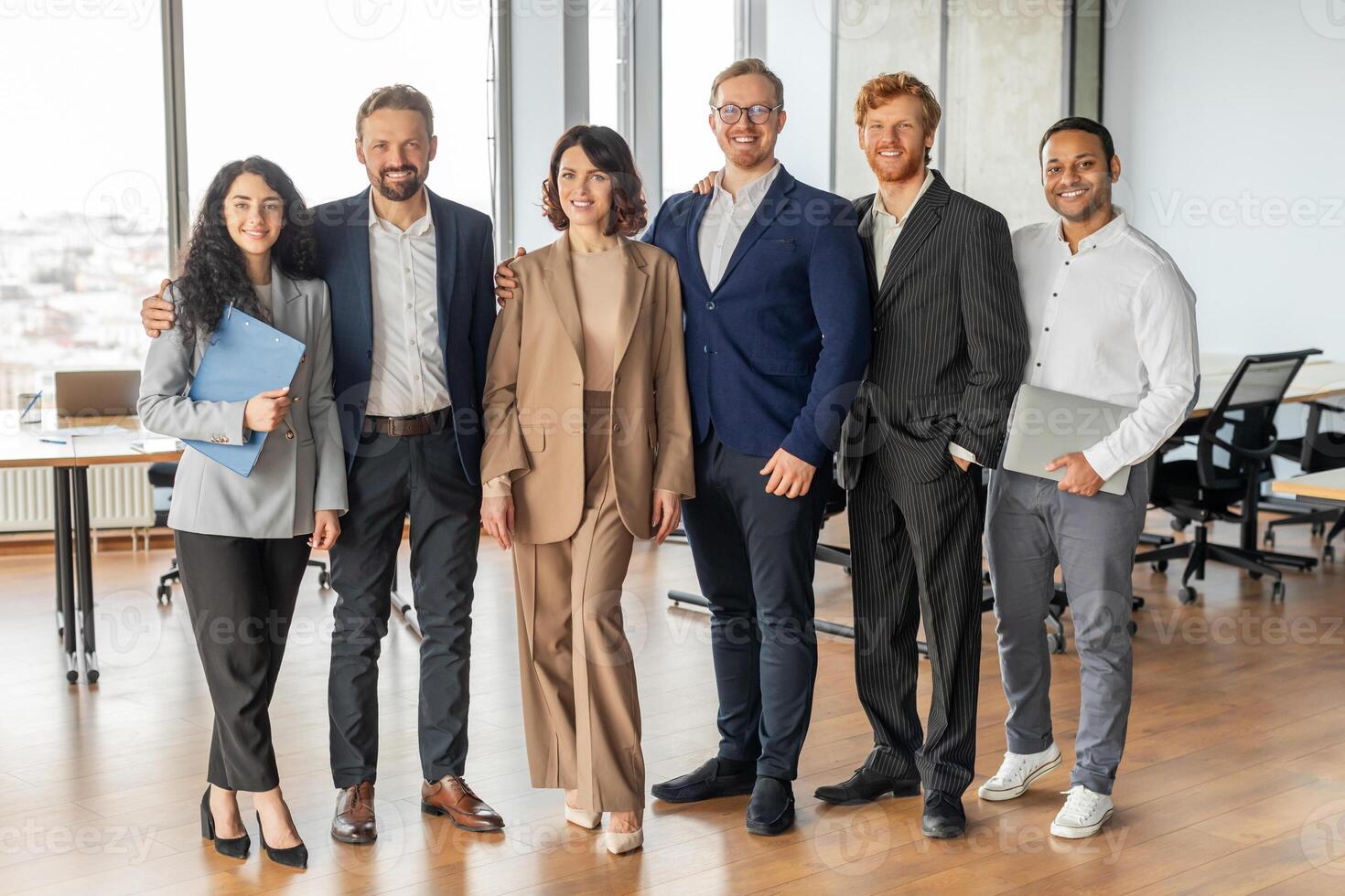 A group of six colleagues stand in a modern office, smiling, looking at the camera. They are a diverse group of professionals in business attire, demonstrating a company culture that values inclusion. photo