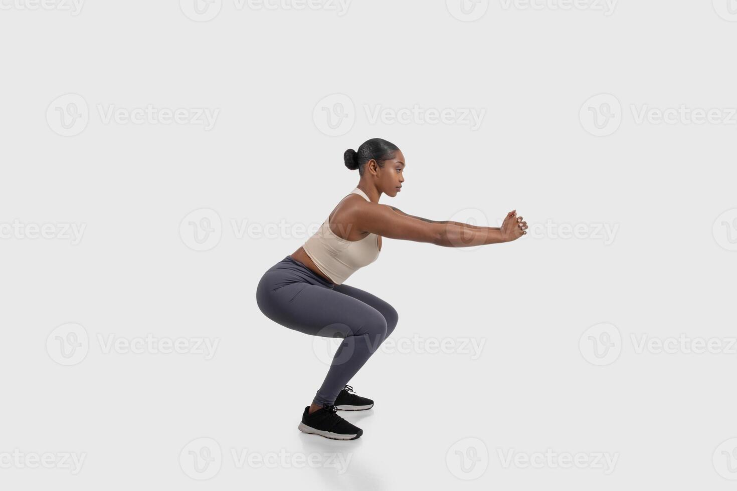 African American woman is seen on a white background, demonstrating a squat exercise. Her body is positioned with feet shoulder-width apart, side view photo