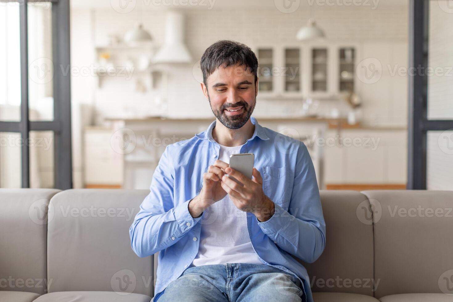 A man wearing a light blue shirt and jeans sits on a couch in a living room, smiling as he uses a smartphone. There are white cabinets in the background and a large window photo