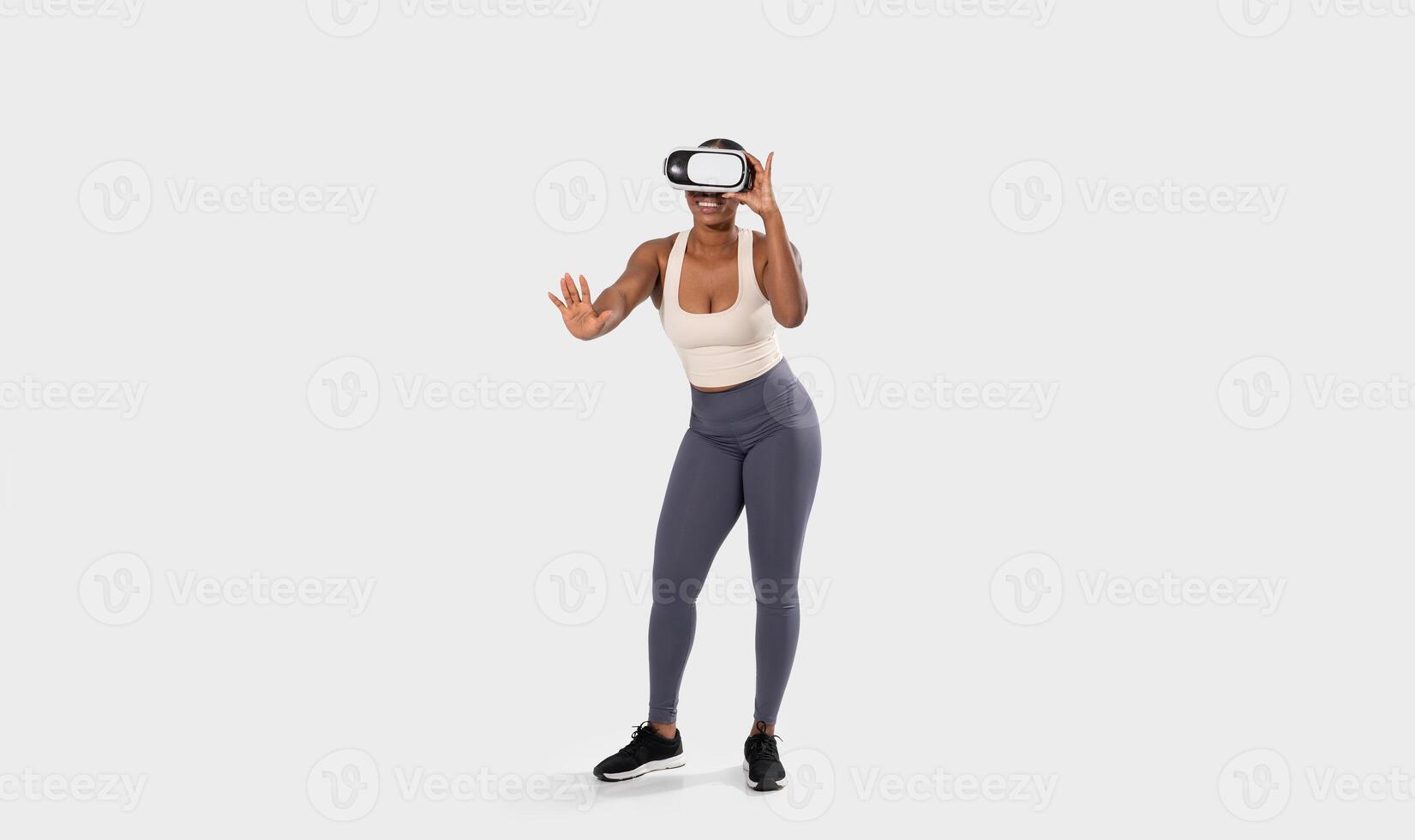 African American woman in athletic clothing and a virtual reality headset is interacting with the virtual world. Her hand is extended, and her expression indicates she is engaged in the experience. photo