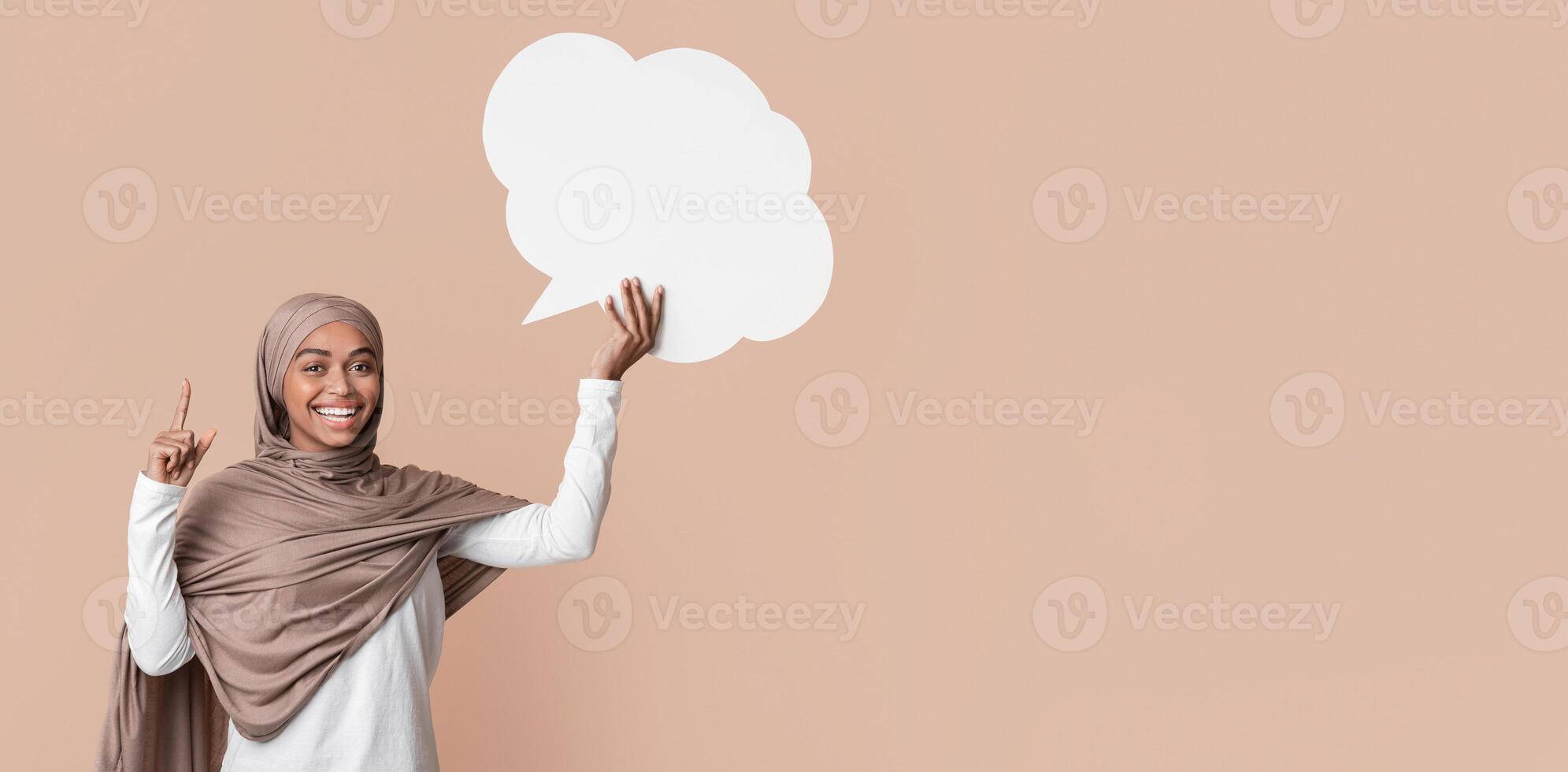 Idea Concept. Joyful black muslim woman in hijab holding empty speech bubble and pointing index finger up over yellow background, copy space photo