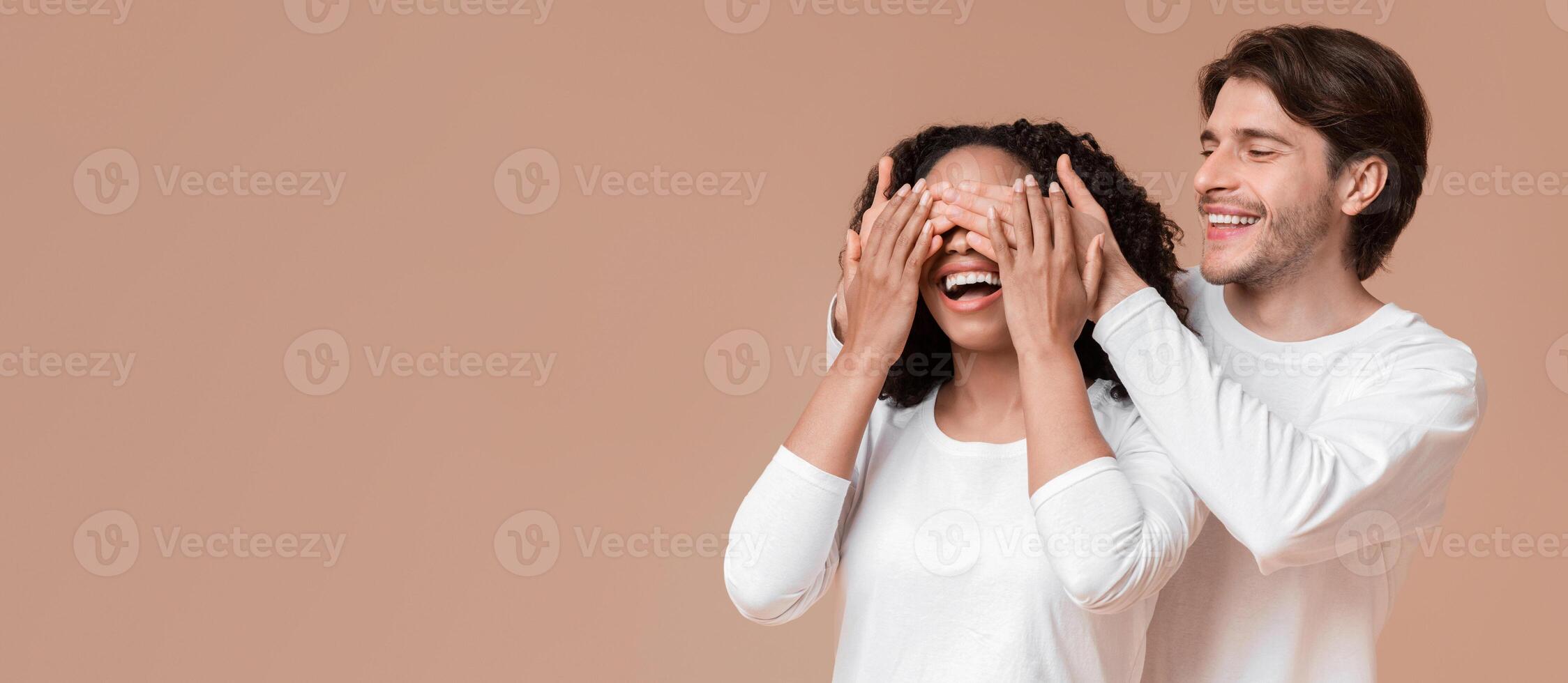 Guess who. Loving guy surprising his afro girlfriend and covering her eyes with hands from behind, yellow studio background with copy space photo