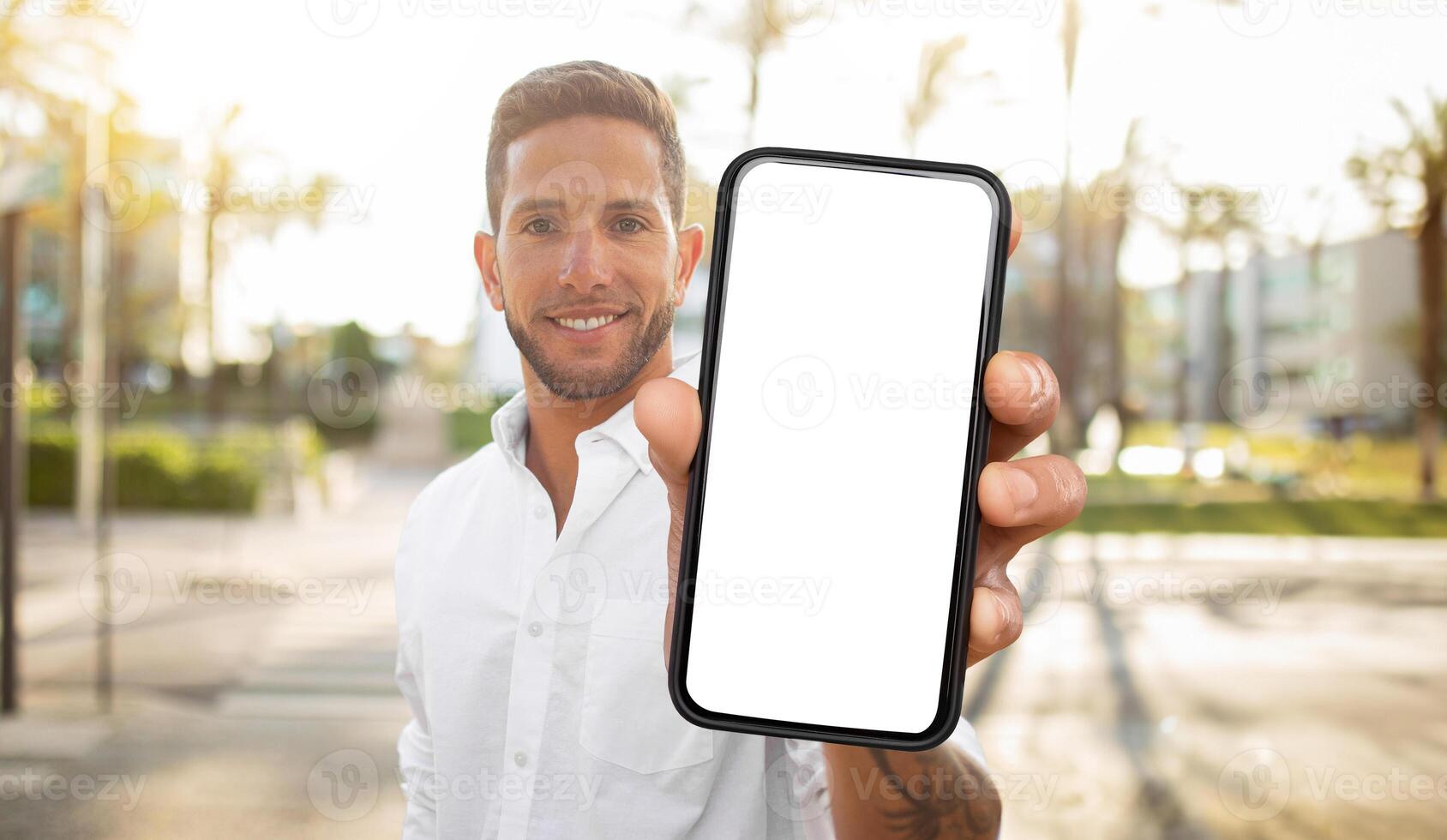 A man in a white shirt stands outdoors, holding a smartphone in front of him with a blank white screen. He is smiling and looking directly at the camera. photo