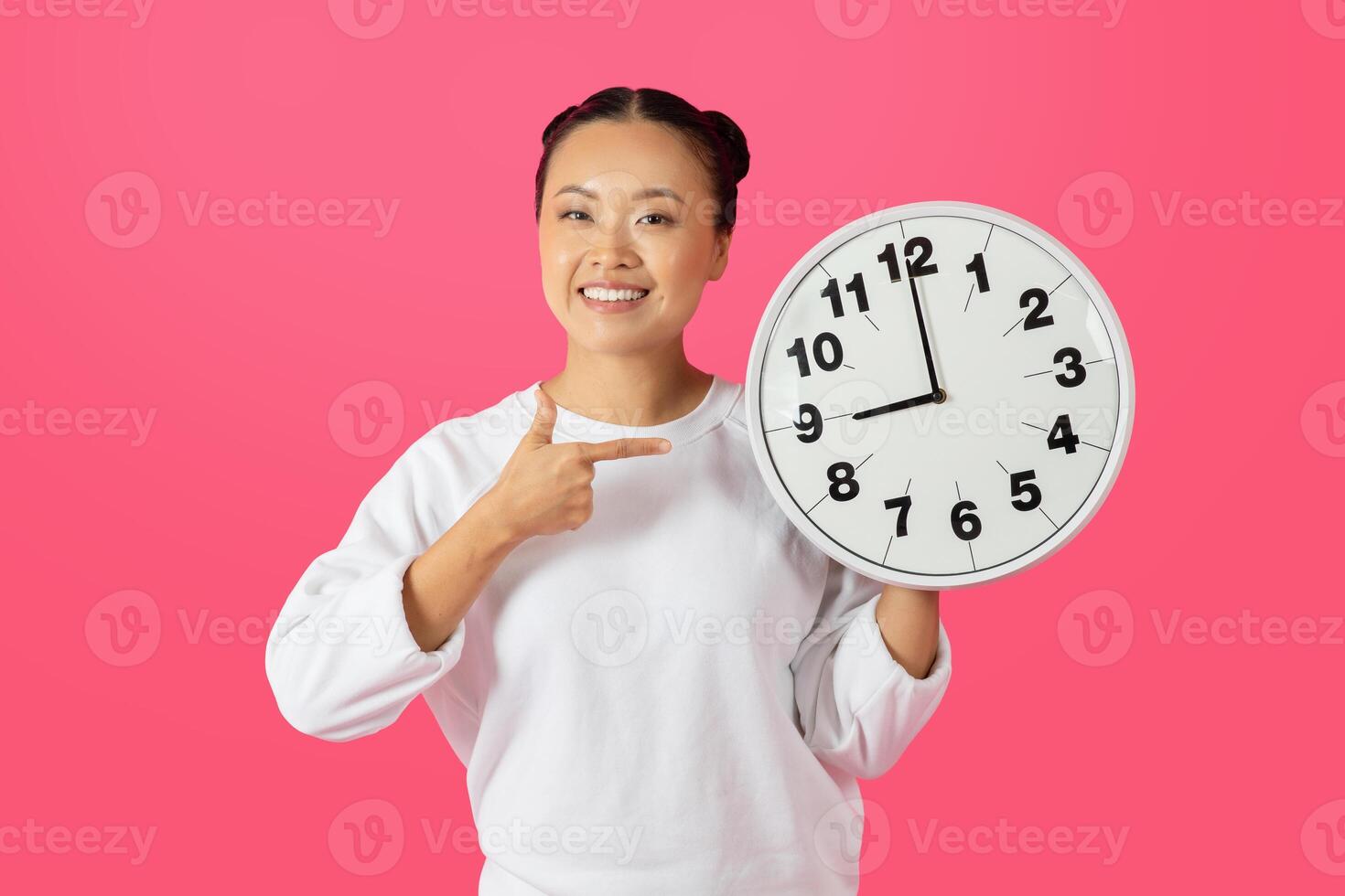 Asian woman with a bright smile holds a white clock in front of her on a pink background. She is wearing a white sweater and pointing at the clock with her right hand. photo