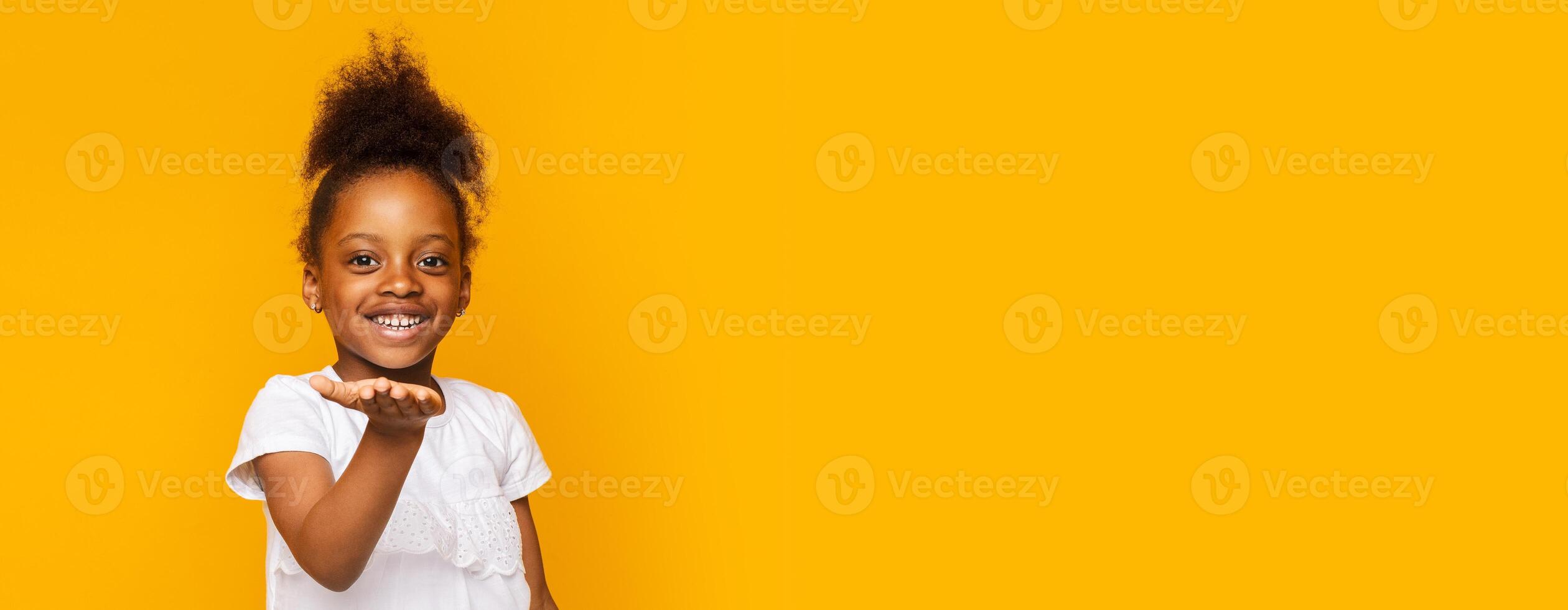 A cheerful young black girl stands confidently, smiling widely as she extends her hand forward. The vibrant yellow backdrop enhances her joyful expression, copy space photo