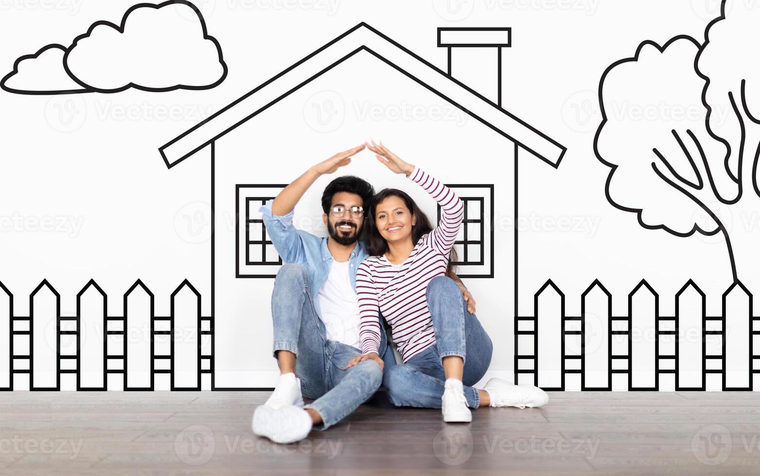 Loving eastern family husband and wife sitting over drawn on white wall house, make a roof with hands and smiling. Real estate, mortgage concept photo
