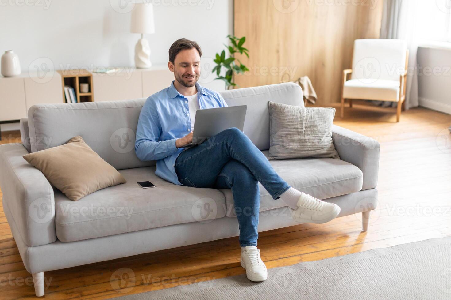 A man is sitting comfortably on a couch, focused on his laptop screen. He appears engaged in typing or browsing online content. The room is well-lit, with various items around him. photo
