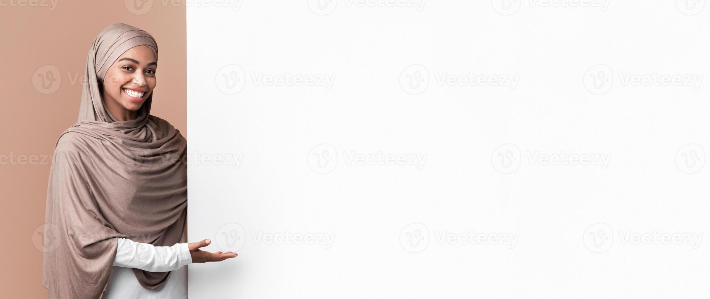 Look here. Smiling black muslim girl in hijab pointing at white advertisement board for your text with open palm, yellow studio background photo