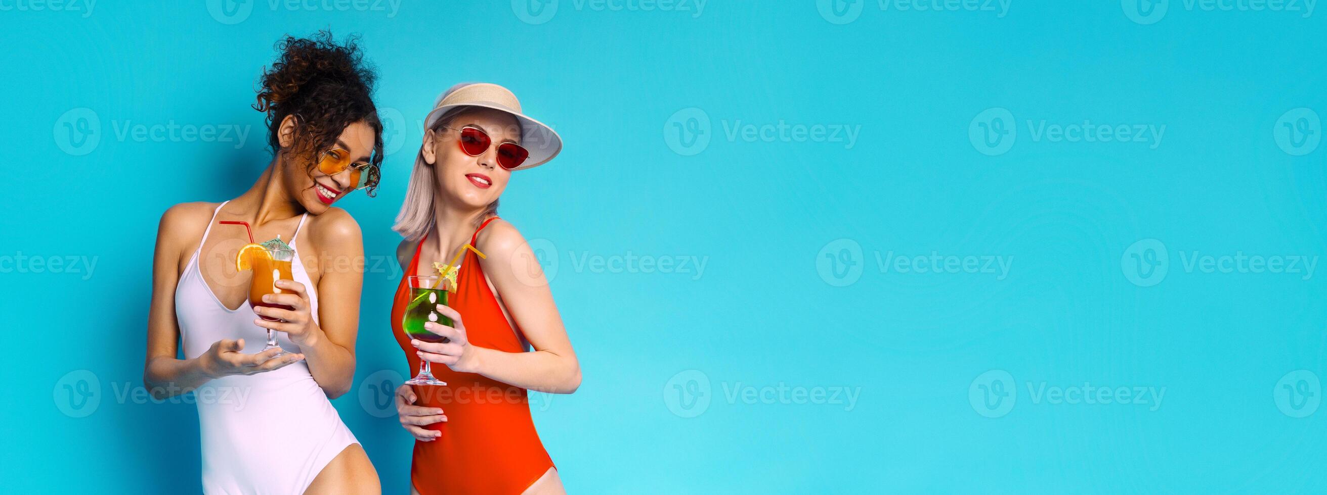 Two women wearing swimsuits and sunglasses are smiling and holding cocktails in front of a bright blue background. They appear to be enjoying themselves on a summer day, copy space photo