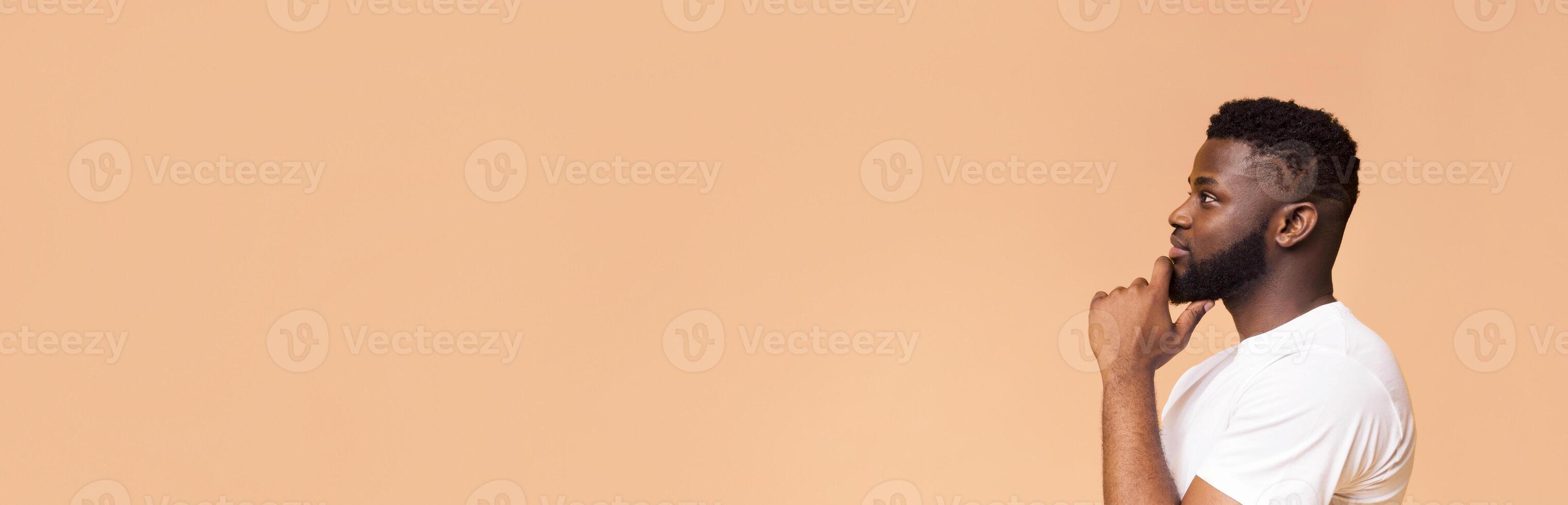 This image features a young African American man thinking while looking to the side. He is wearing a white t-shirt and standing against a peach-colored background, copy space photo