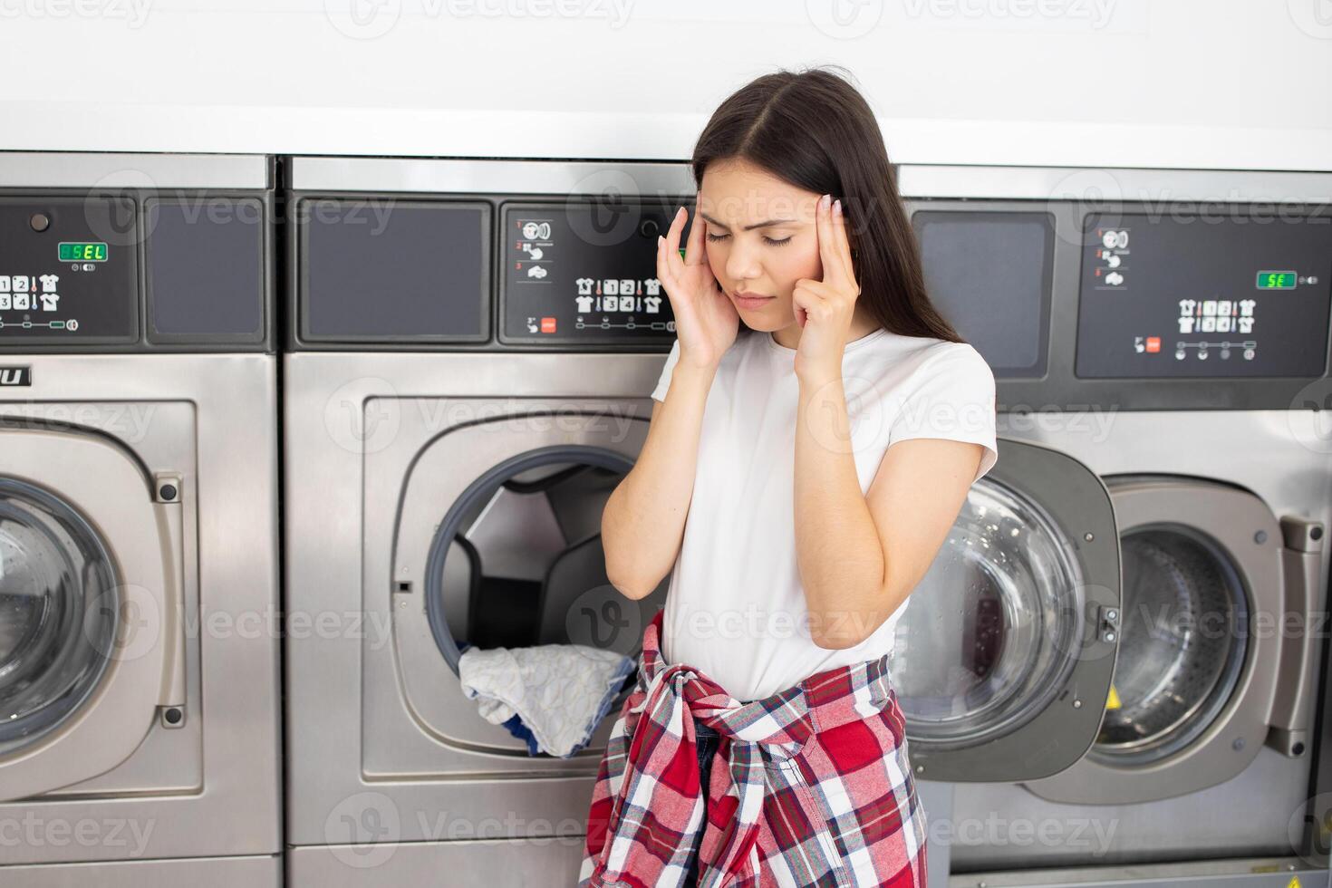 A young woman stands in a laundromat with her hands on her temples, looking stressed. She is dealing with unexpected laundry issues while several machines operate in the background. photo