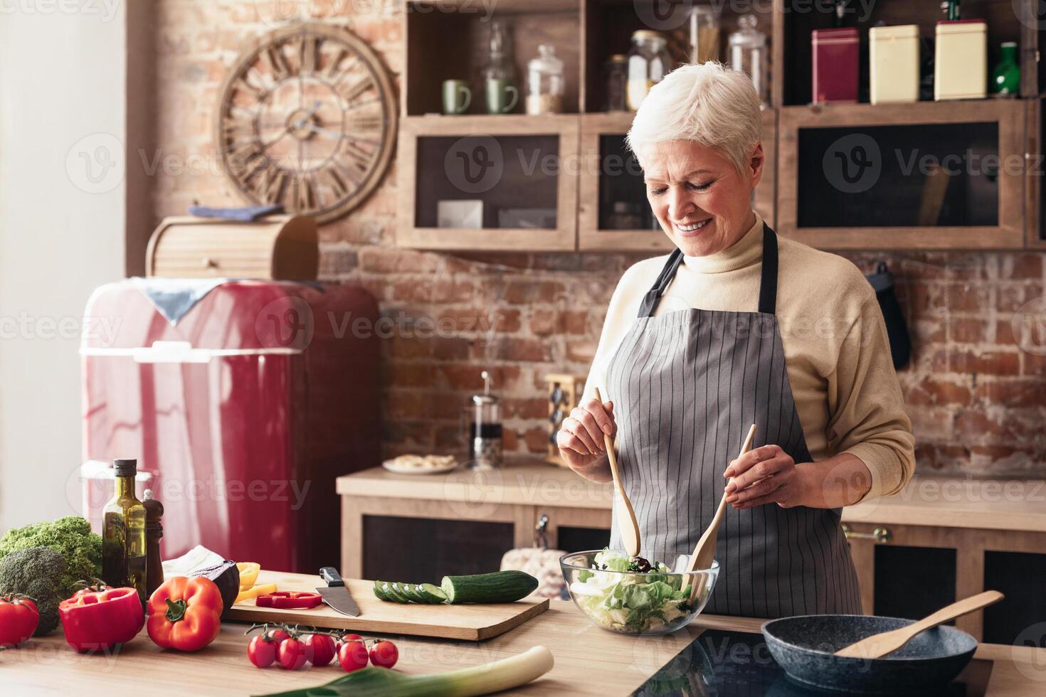 A senior woman in an apron smiles as she uses wooden spoons to mix a salad in a glass bowl on a kitchen counter. A red refrigerator is visible in the background, and other ingredients photo
