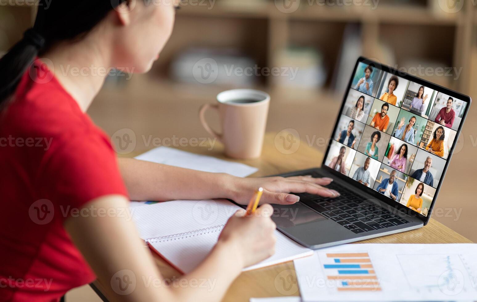 A woman sits at a desk with a laptop open in front of her. She is taking notes in a notebook with a pencil while attending an online meeting. The laptop screen displays a conference photo