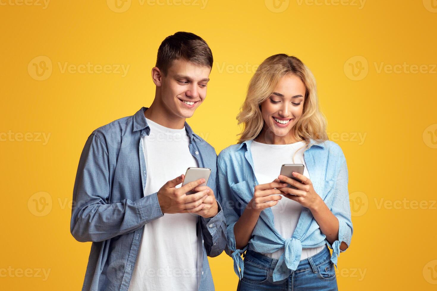 Curious boyfriend peeking into girlfriend's smartphone, trying to read her messages, yellow studio background with copy space photo