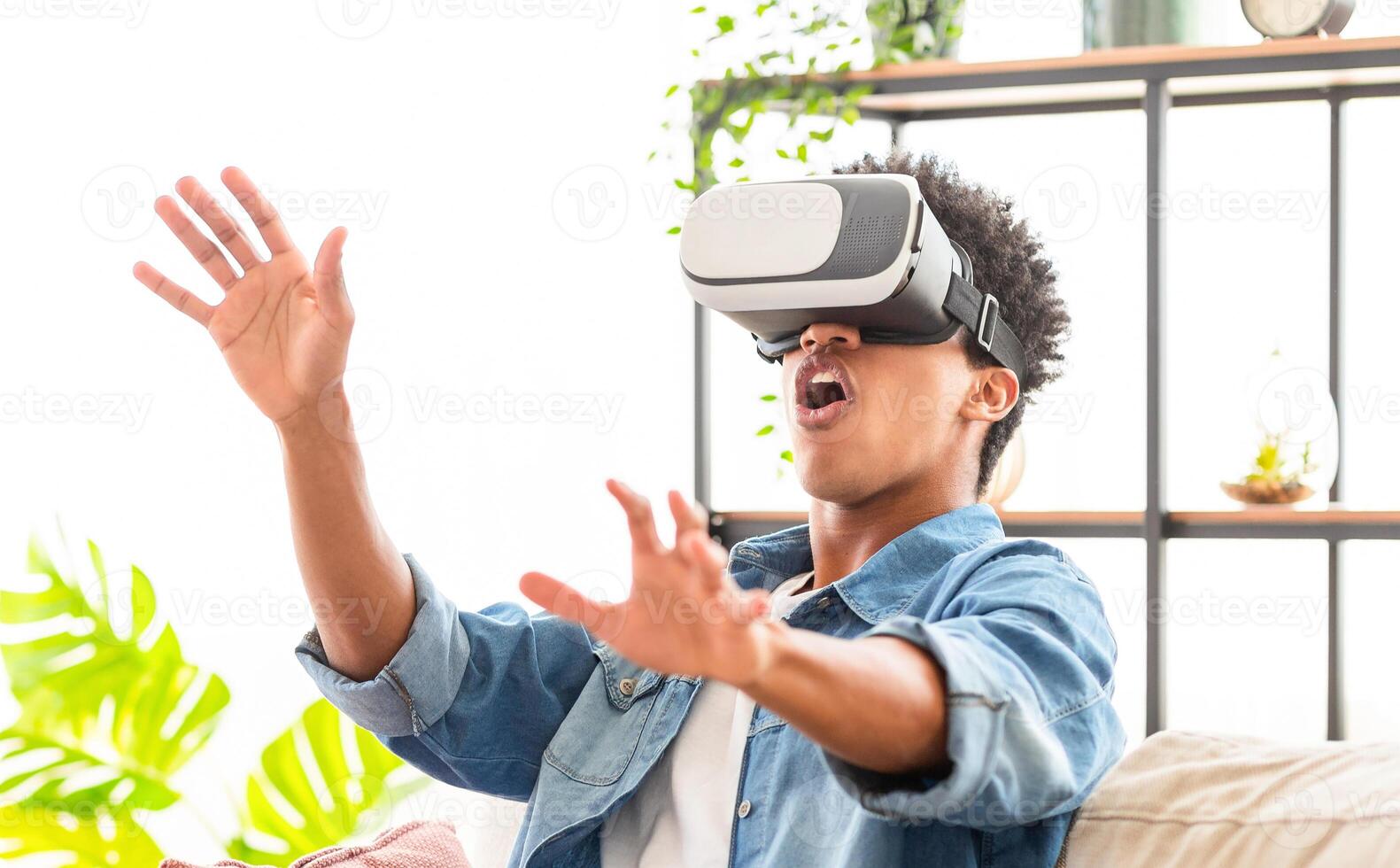 African American teen guy wearing a virtual reality headset sits on a couch, his hands outstretched, and his mouth open in surprise. He appears to be immersed in a virtual reality experience photo