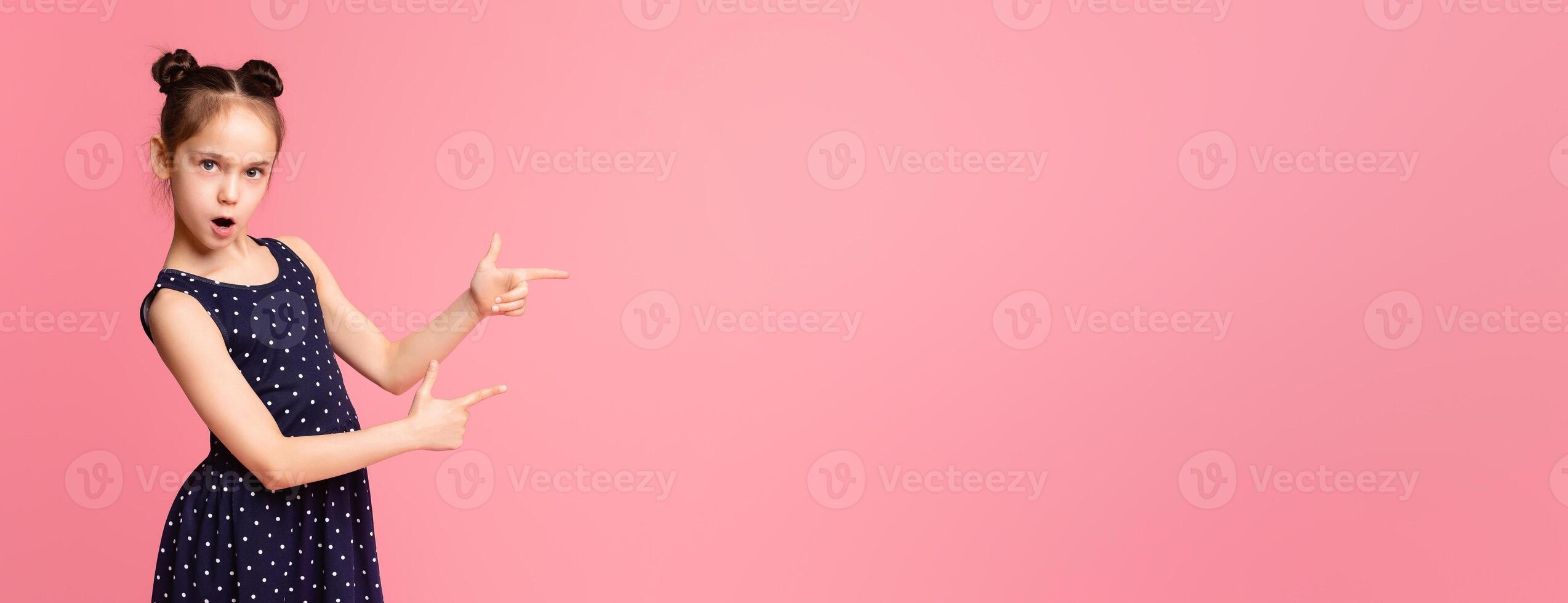 A young girl wearing a blue dress is pointing at an object off-camera, her expression curious and engaged, hair is tied back in a ponytail, and she stands in front of a neutral background, copy space photo