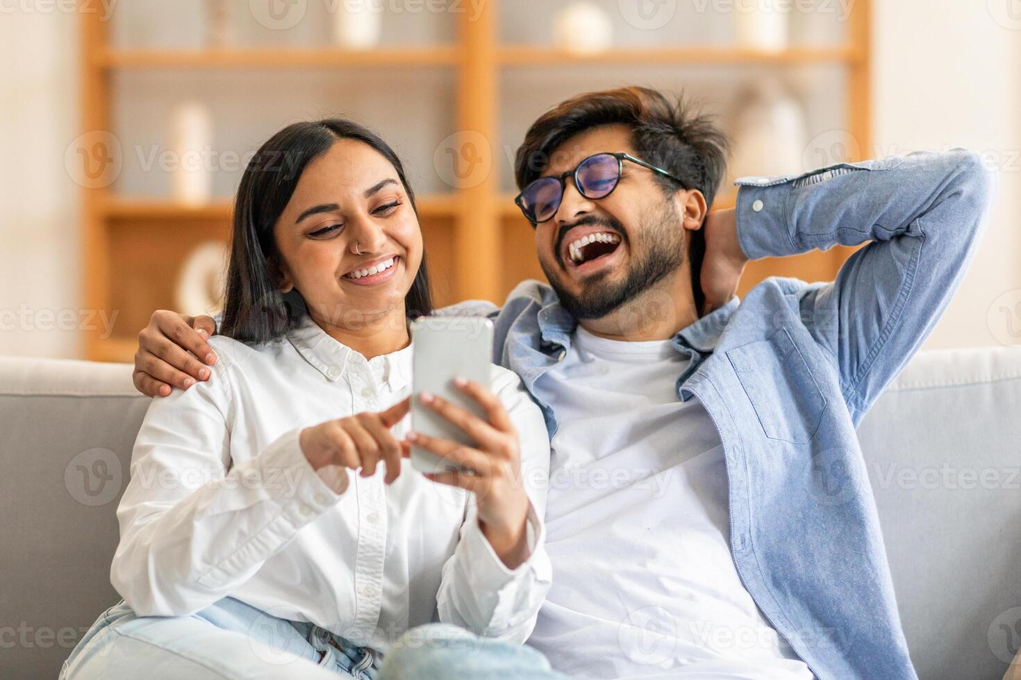 Indian man and woman are seated on a couch, their attention focused on a cell phone held between them. Both individuals appear engaged in the screens content, immersed in digital interaction. photo