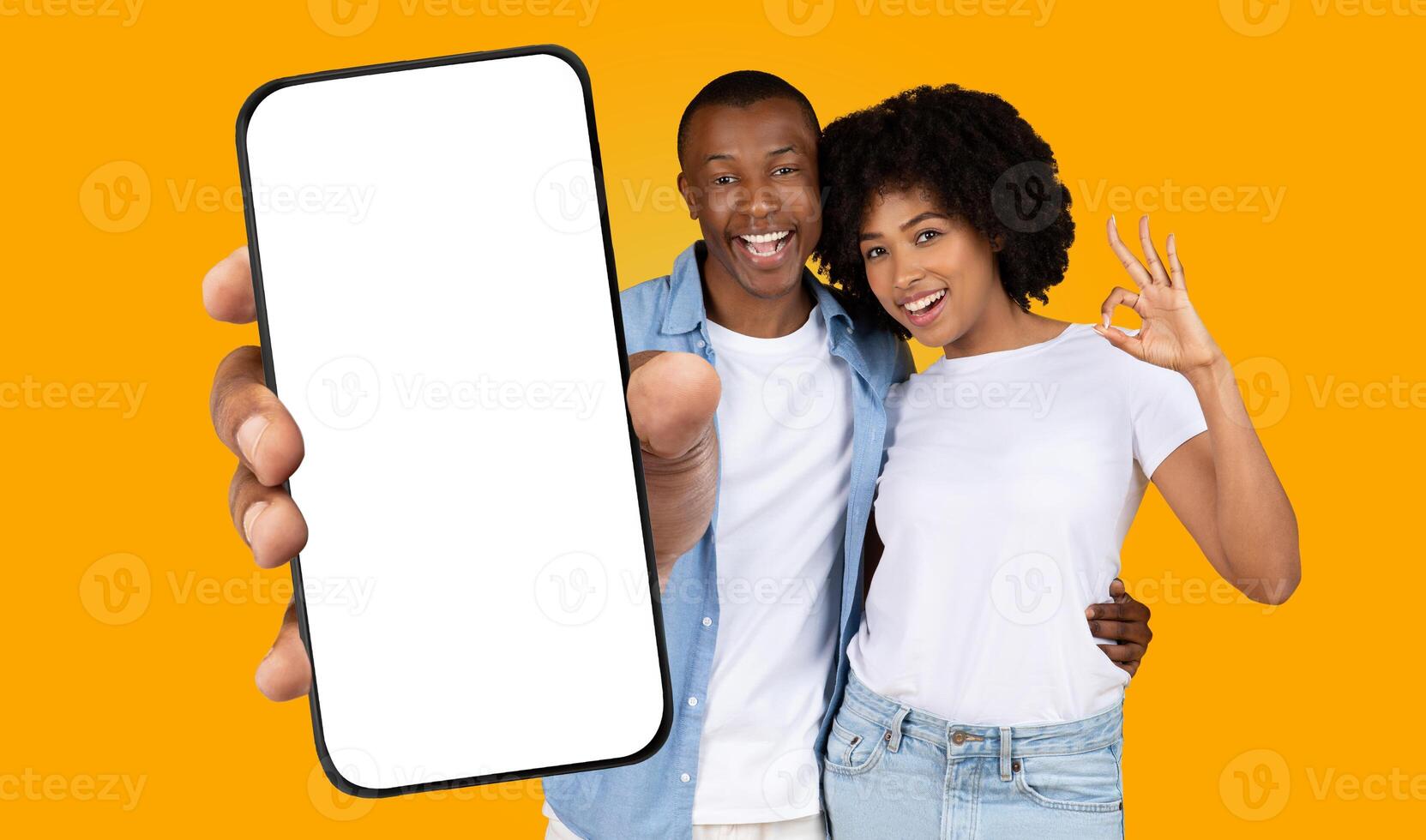 Black man and woman stand in front of yellow background. The man is holding a smartphone with a blank screen. The woman is making the ok sign with her hand. They both have big smiles on their faces. photo