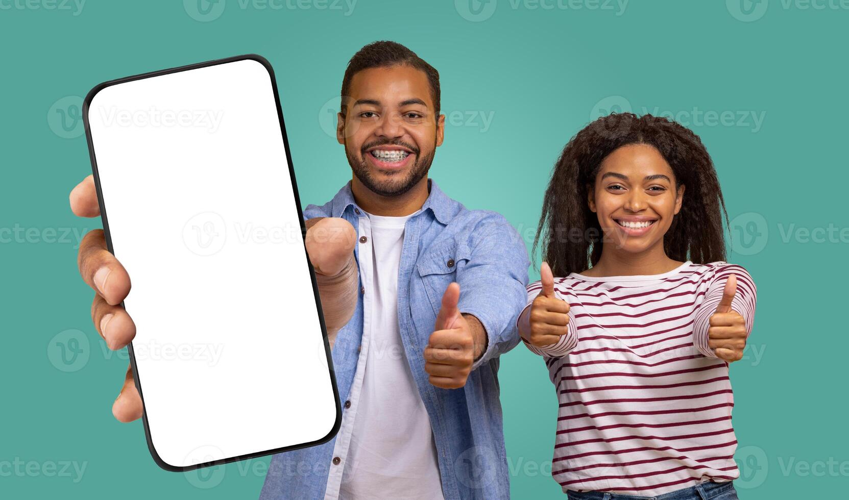 A smiling black man and woman stand in front of a teal background. The man is holding a smartphone with a blank screen up to the camera. He and the woman both have their thumbs up photo