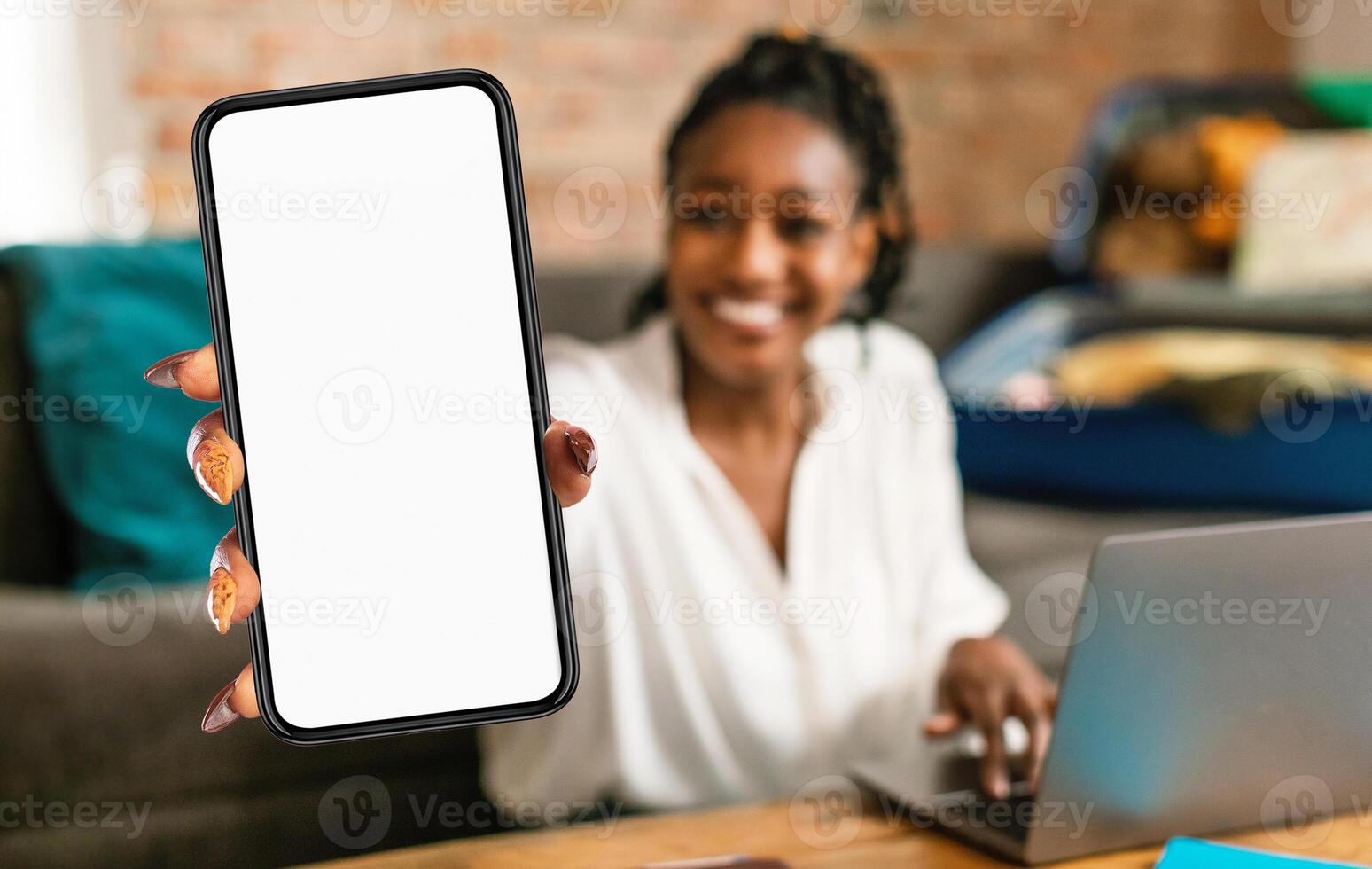 Black woman is seen holding up a cell phone in front of a laptop screen. She appears to be engaged in a call or browsing content on the phone while using the laptop simultaneously. photo