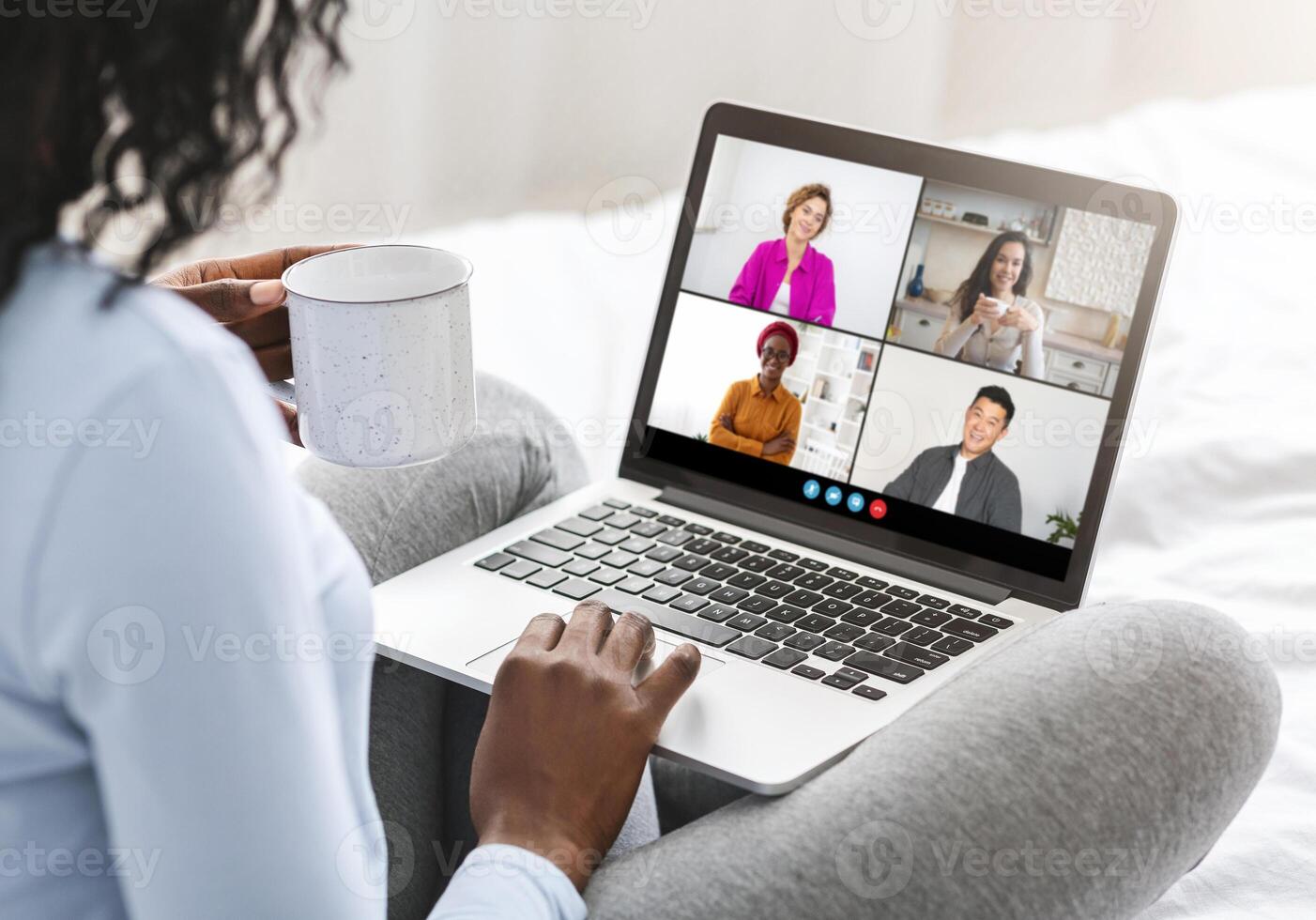 African American woman sits in bed with a laptop on her lap, holding a mug. On the screen is a call with four other people, suggesting a work meeting or social gathering. photo