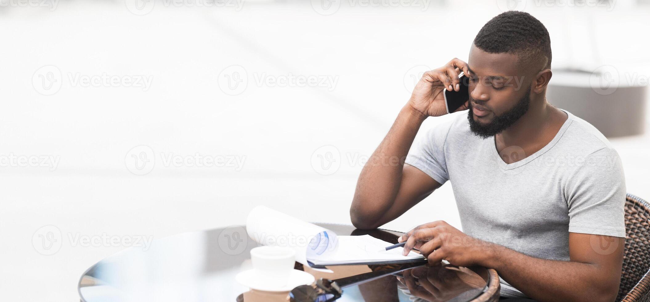A man sits at a table, taking notes while on a phone call. Hes looking at a document, wearing a light grey t-shirt, and has a short beard. photo