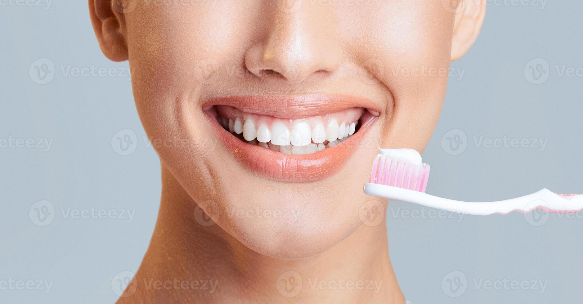 A close-up of a woman face as she smiles, with a toothbrush and toothpaste held near her mouth. The image focuses on her bright white teeth and happy expression. photo