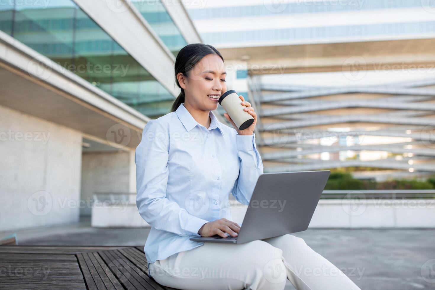 A young Asian woman sits comfortably on a wooden bench, sipping coffee from a cup while focused on her laptop. The contemporary architecture and bright surroundings create a vibrant atmosphere photo