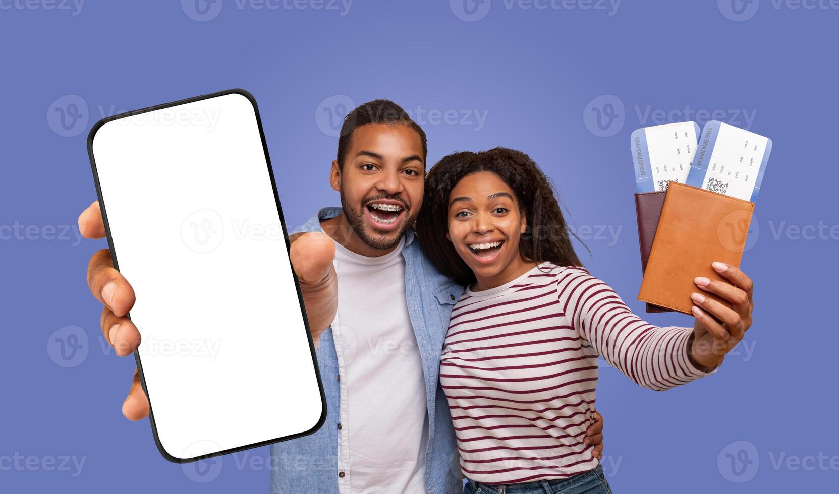 A young black couple smiling excitedly at the camera while holding a blank smartphone and boarding passes. The man is pointing at the phone, and the woman is holding a passport holder with tickets. photo