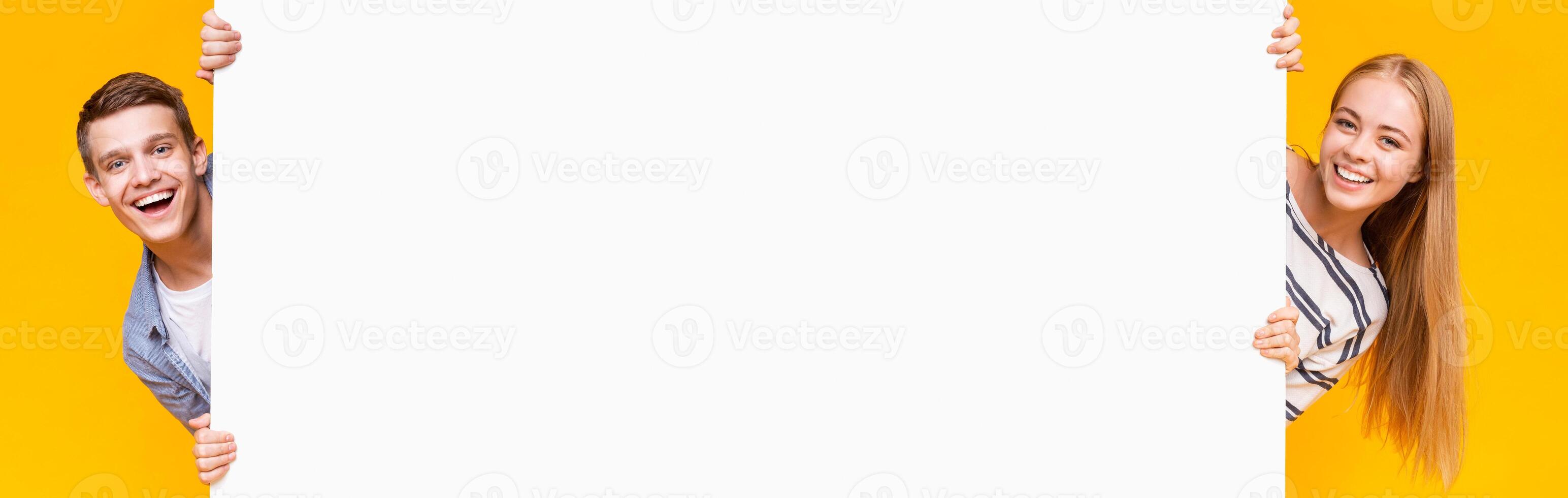 Two young multiracial guys standing behind long empty board for advertising over yellow background photo
