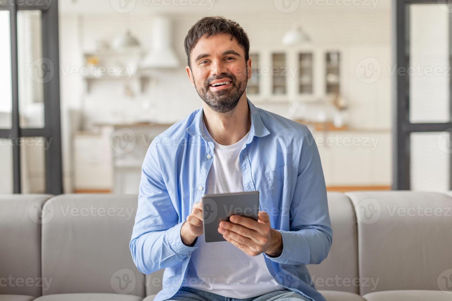 A man sits on a gray couch in his home and smiles while holding a tablet computer in his hands, websurfing or scrolling photo