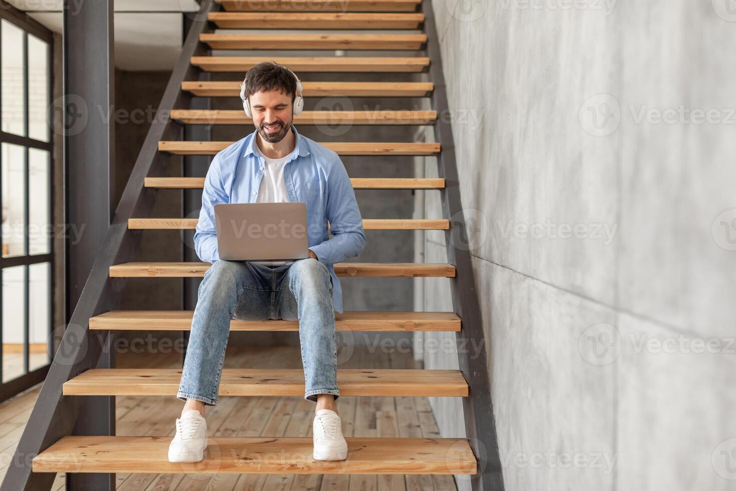 A man sits on wooden stairs in a modern home. He is wearing a blue shirt and jeans and is looking down at a laptop in his lap. He is smiling and has headphones on. He appears to be working photo