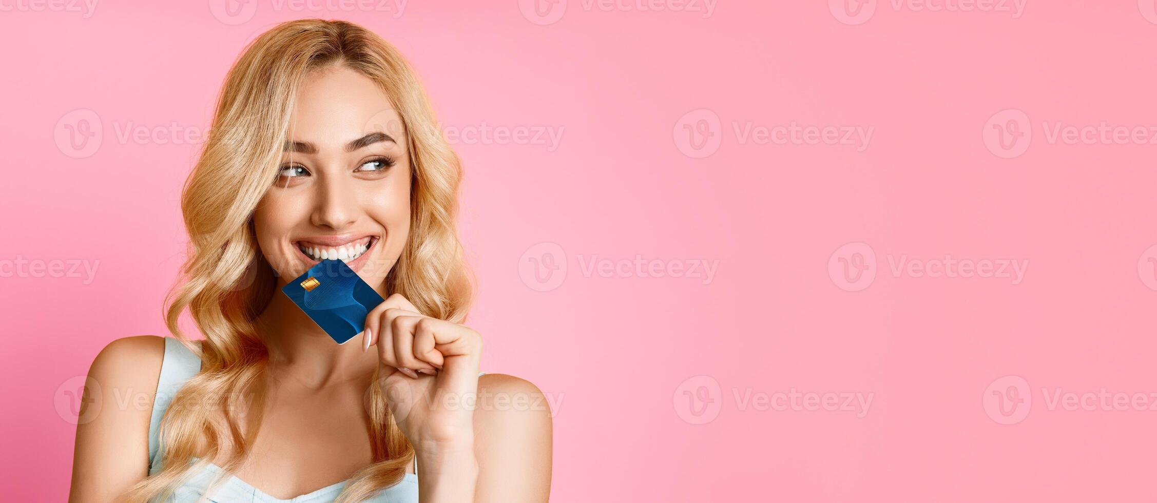 A young woman with blonde hair smiles brightly while holding a blue credit card in her mouth and looking off to the side against a plain pink background, panorama with copy space photo