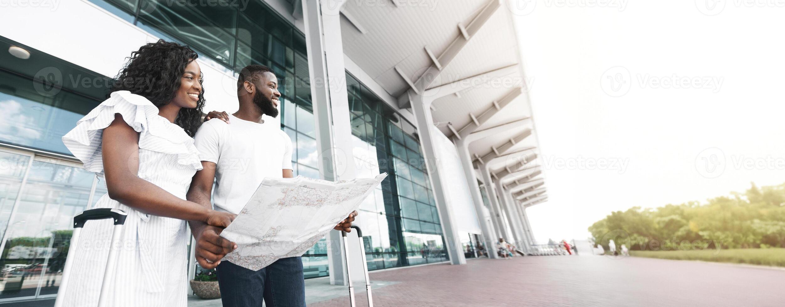 Black couple, dressed in casual clothing, stand outside a modern airport terminal. They are looking at a map, presumably trying to find their way around, copy space photo