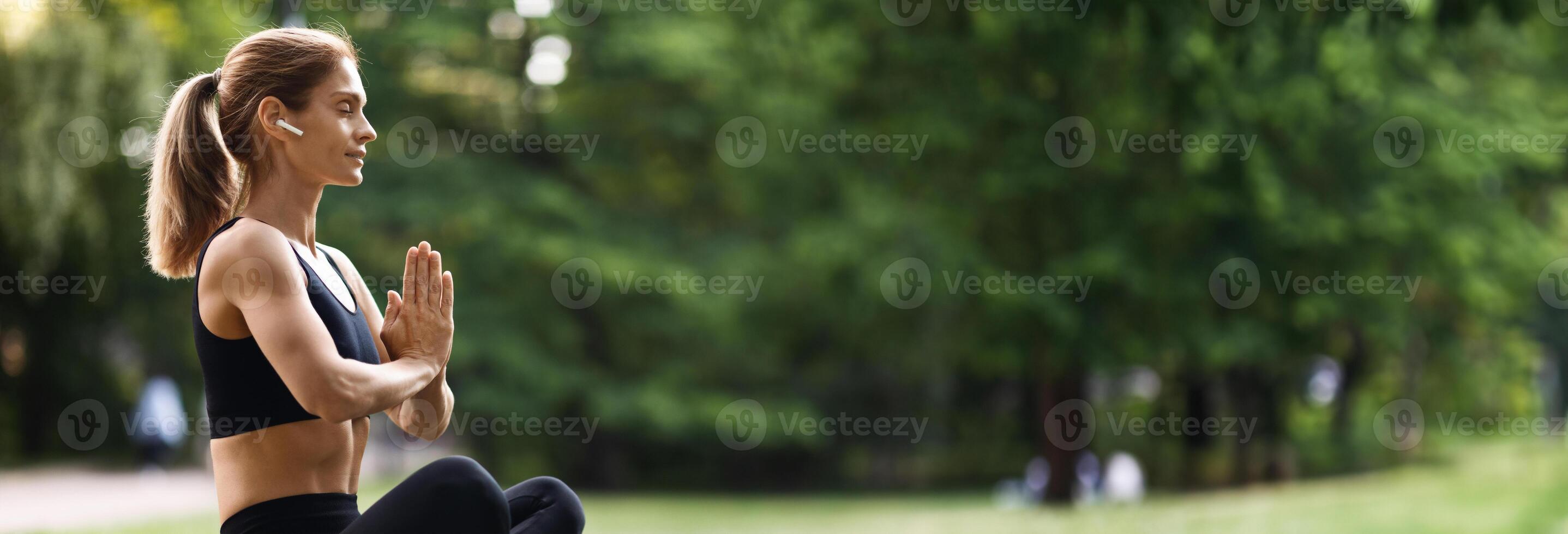 Peaceful millennial blonde woman in black tight sportswear sitting on fitness mat in lotus position with closed eyes, using wireless earbuds, meditating alone at public park, side view, copy space photo