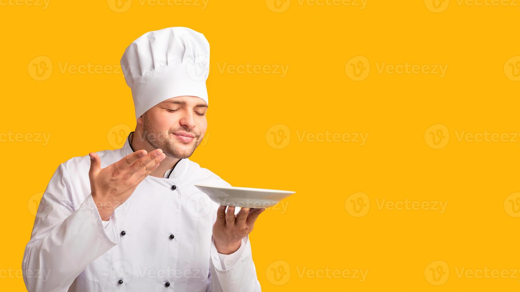 Professional Male Chef Holding Empty Plate Smelling The Aroma Of Invisible Dish Over Yellow Studio Background. Panorama photo