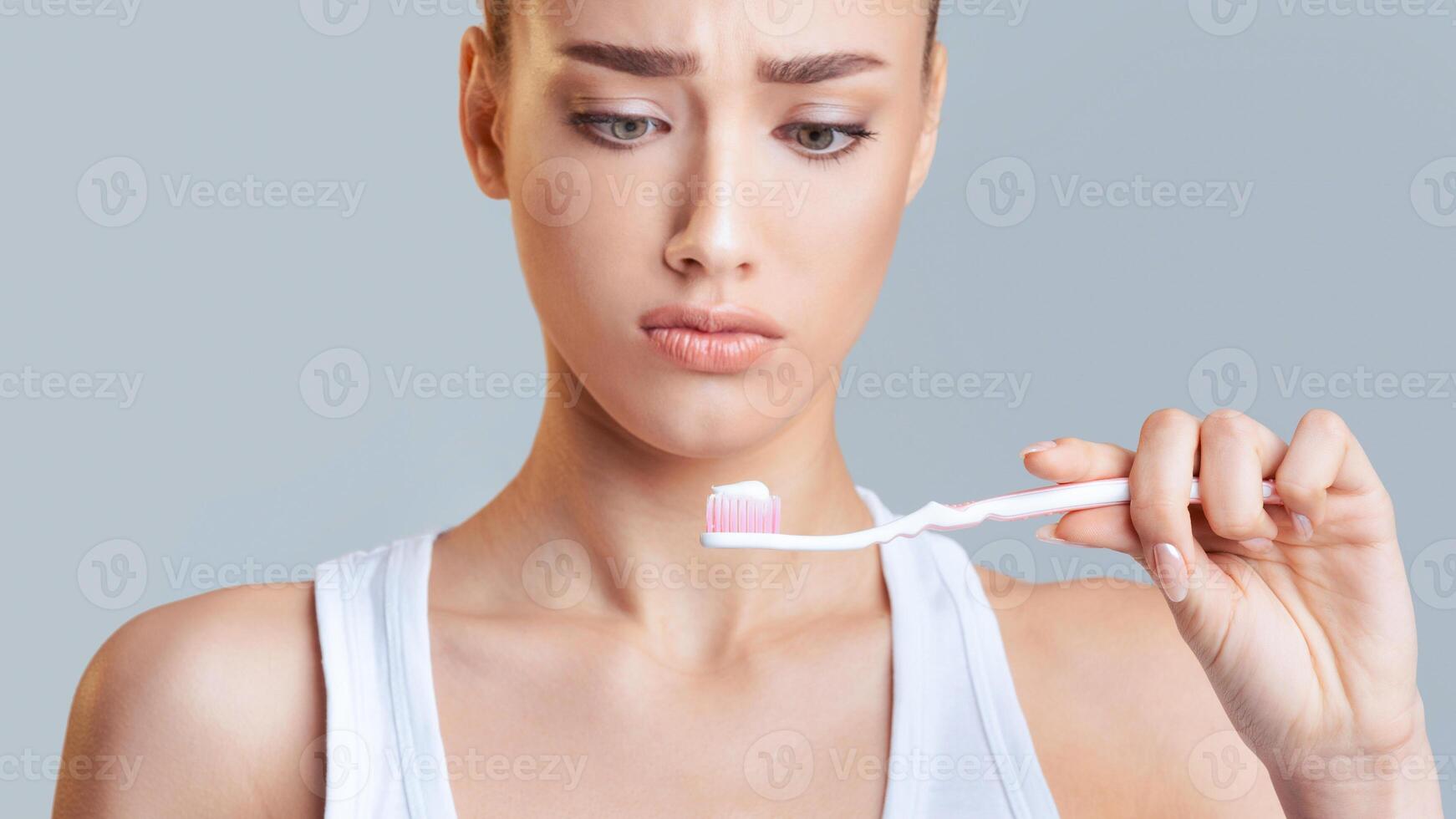 The image features a close-up portrait of a young woman holding a toothbrush with toothpaste on it. She is looking down at the toothbrush with a slightly worried expression on her face. photo