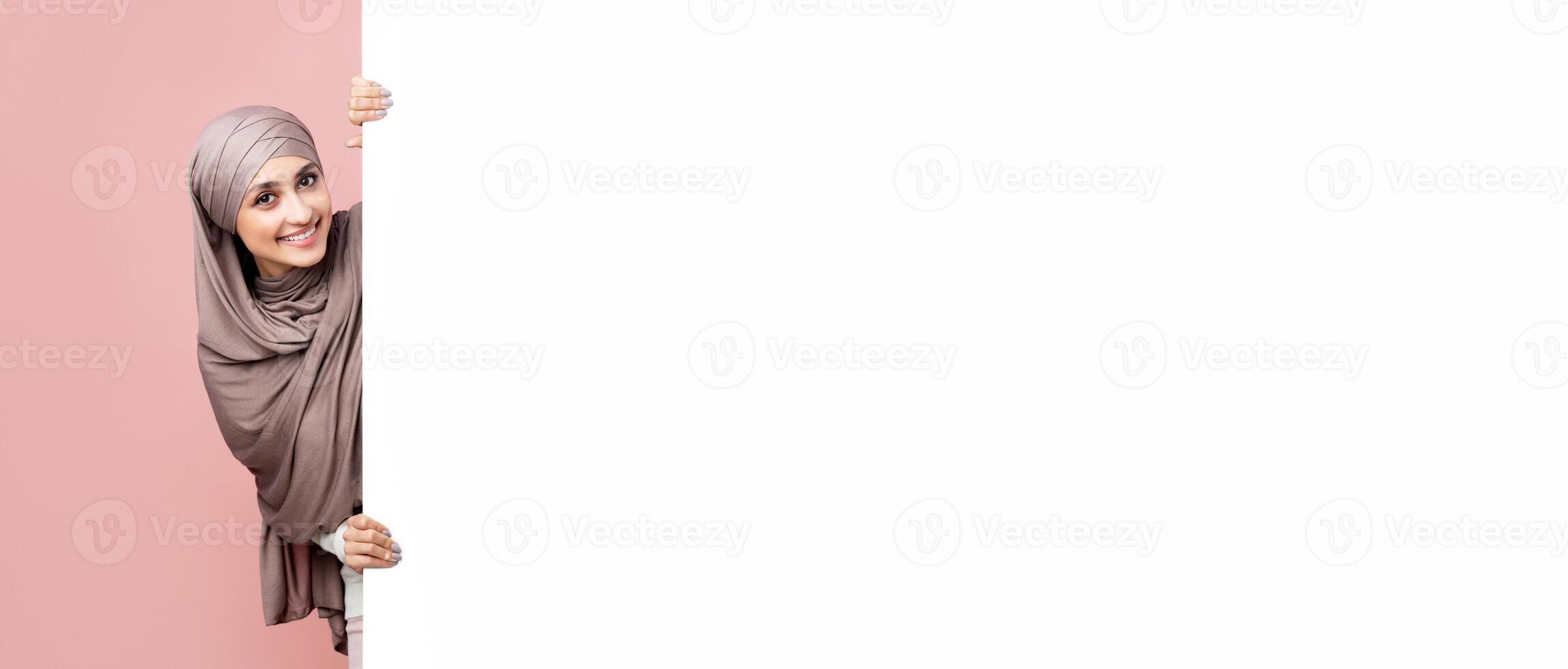 Free space for advertisement. Beautiful muslim girl in hijab standing next to white board for your text or ad, pink studio background photo