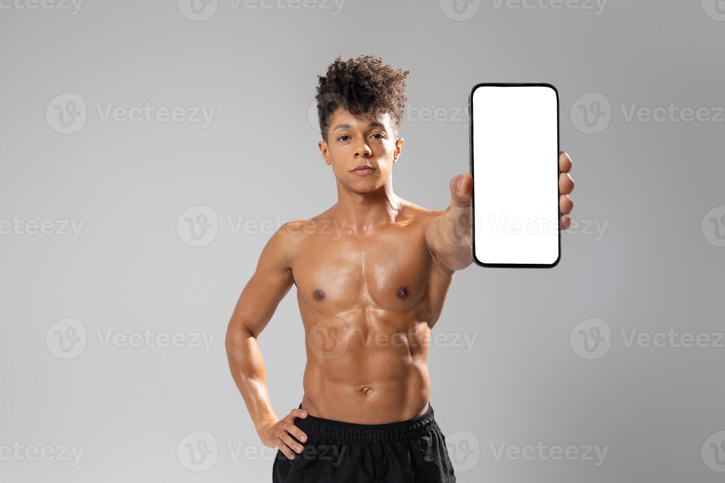 A young man stands confidently with a toned physique, showcasing a blank smartphone to the camera. His athletic stance emphasizes strength, while the neutral background photo