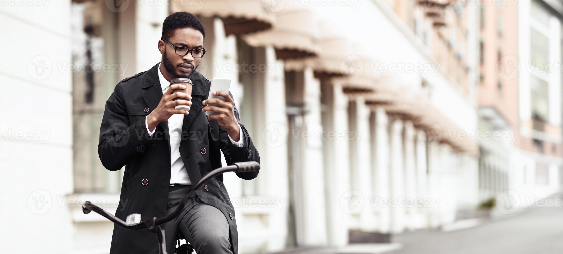 An African American businessman is biking to the office while using his phone. The scene is energetic and urban, highlighting the blend of modern technology and eco-friendly commuting. photo