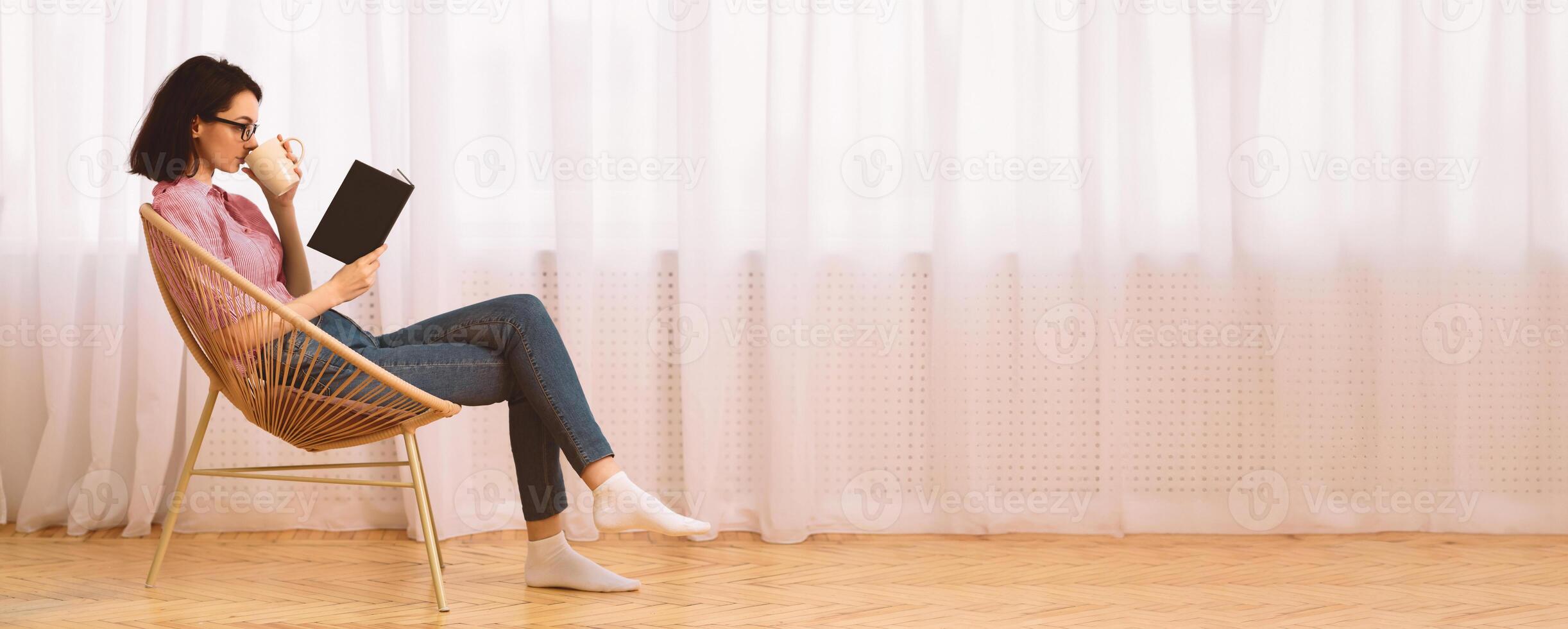 Free Time. Young woman in glasses reading book sitting in chair by the window on spring morning, drinking tea, copyspace photo